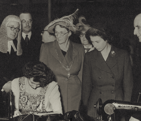 A black and white photograph of HRH Princess Elizabeth visiting Enderley Mills in Newcastle. She looks down on a woman using a singer sewing machine.