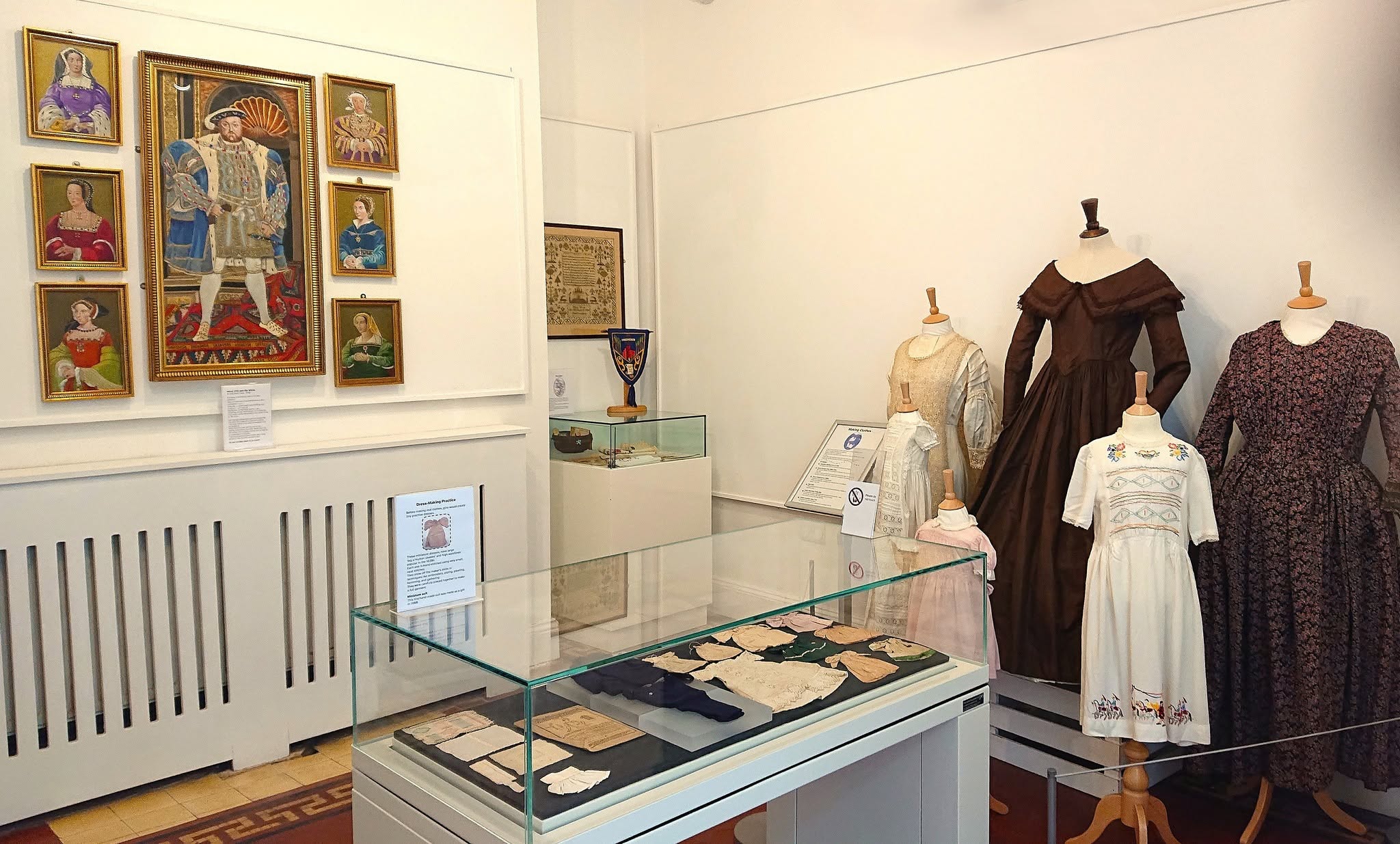 A museum room containing a textile exhibition, with cases of objects, dressed mannequins and on the wall a seven part of embroidery of Henry VIII and this wives