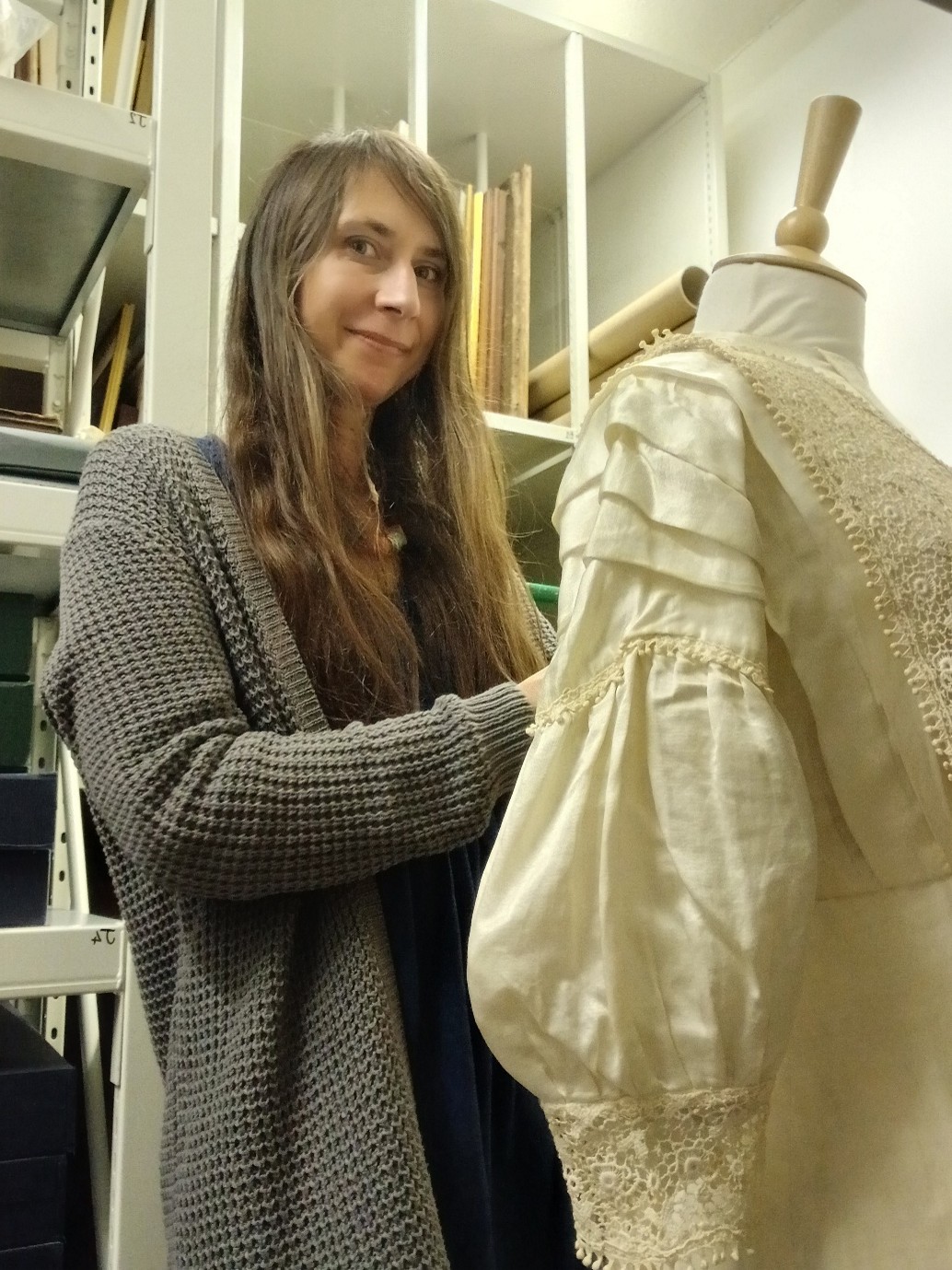 A lady with long brown hair, wearing a grey cardigan is standing in a room with shelves behind her. She has just finished placing an Edwardian cream satin wedding dress on the mannequin in front of her.