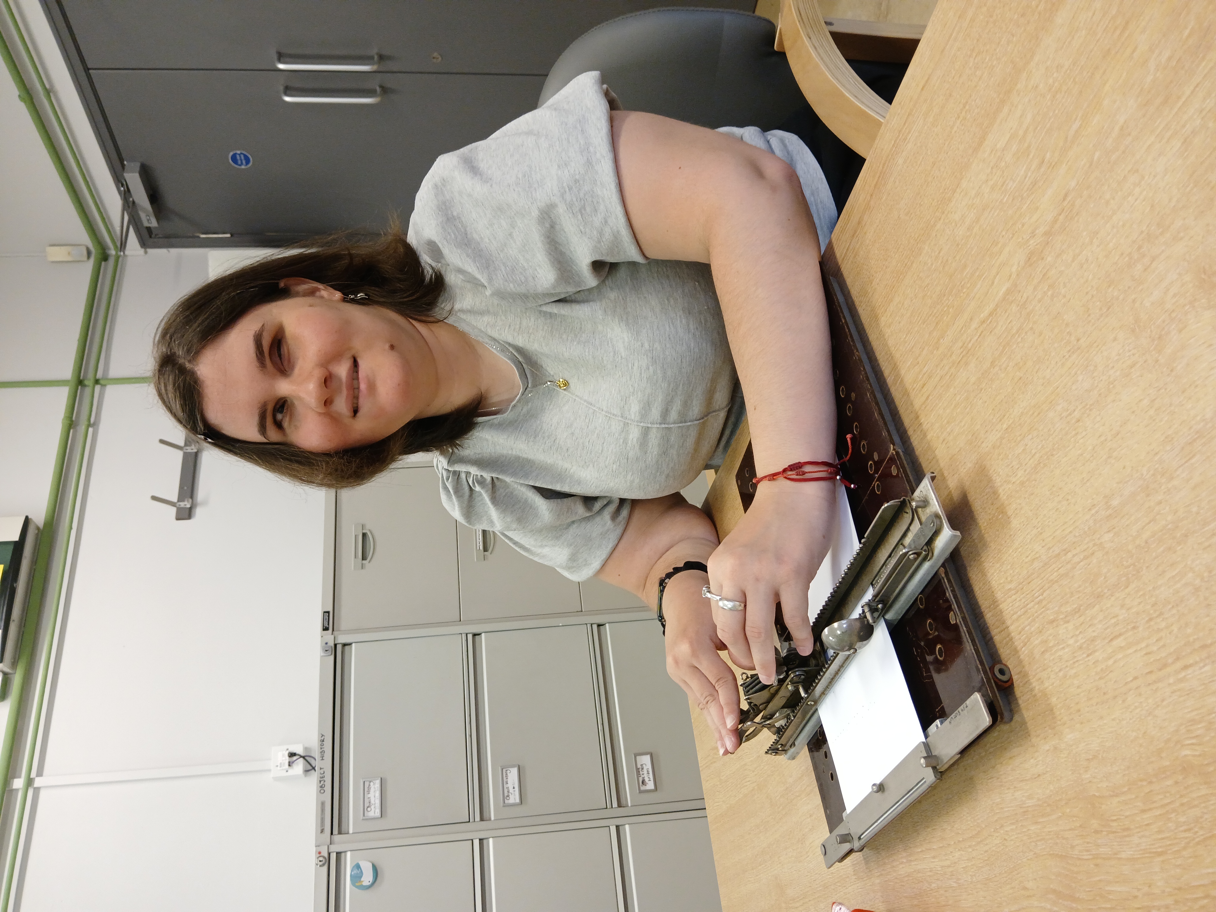 Klaudia young female, with short brown hair, wearing a grey top sits at a table with an early brailler. The brailler is a brown flat rectangle with a piece of white paper attached. A metal part sits on top, Klaudia rests her fingers on the brailler keys.