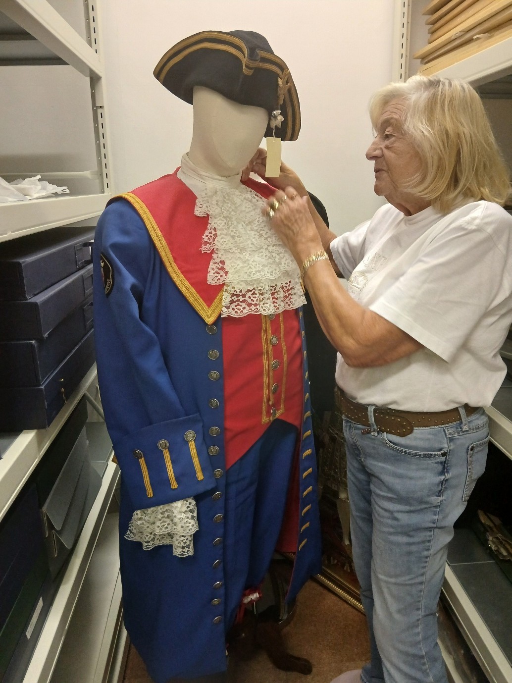 In a shelved storeroom, a woman wearing blue jeans and white t-shirt with short hair stands to the right of a mannequin dressed in an elaborate town crier's red and white outfit, with a frilly white collar and black tricorn hat.