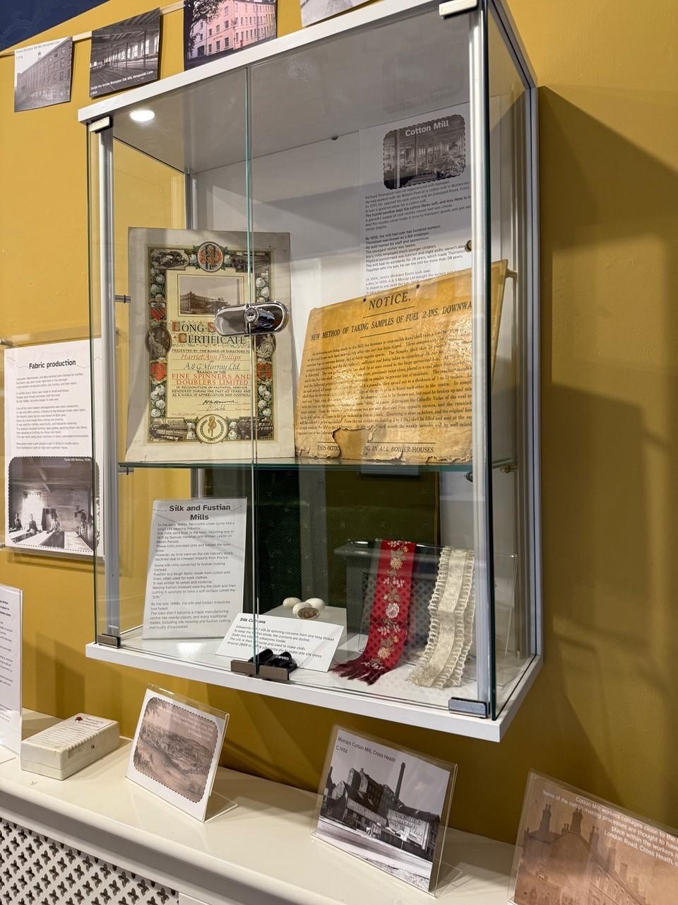 The image shows a display case in the museum Spotlight Gallery with items from the history of textile manufacturing in the borough