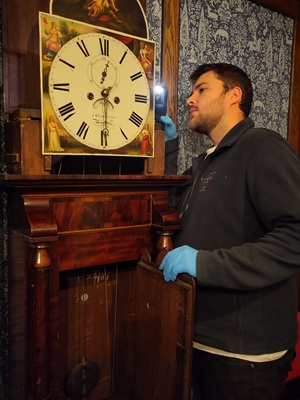 A man in a blue jumper and gloves examines a long case clock with a torch. The front door is open showing the pendulum and the wooden hood is off showing the clock face.