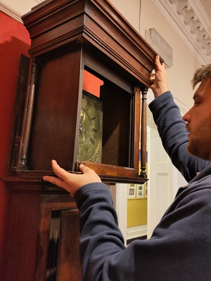 A man in a blue jumper carefully lifts the wooden hood off a long case clock