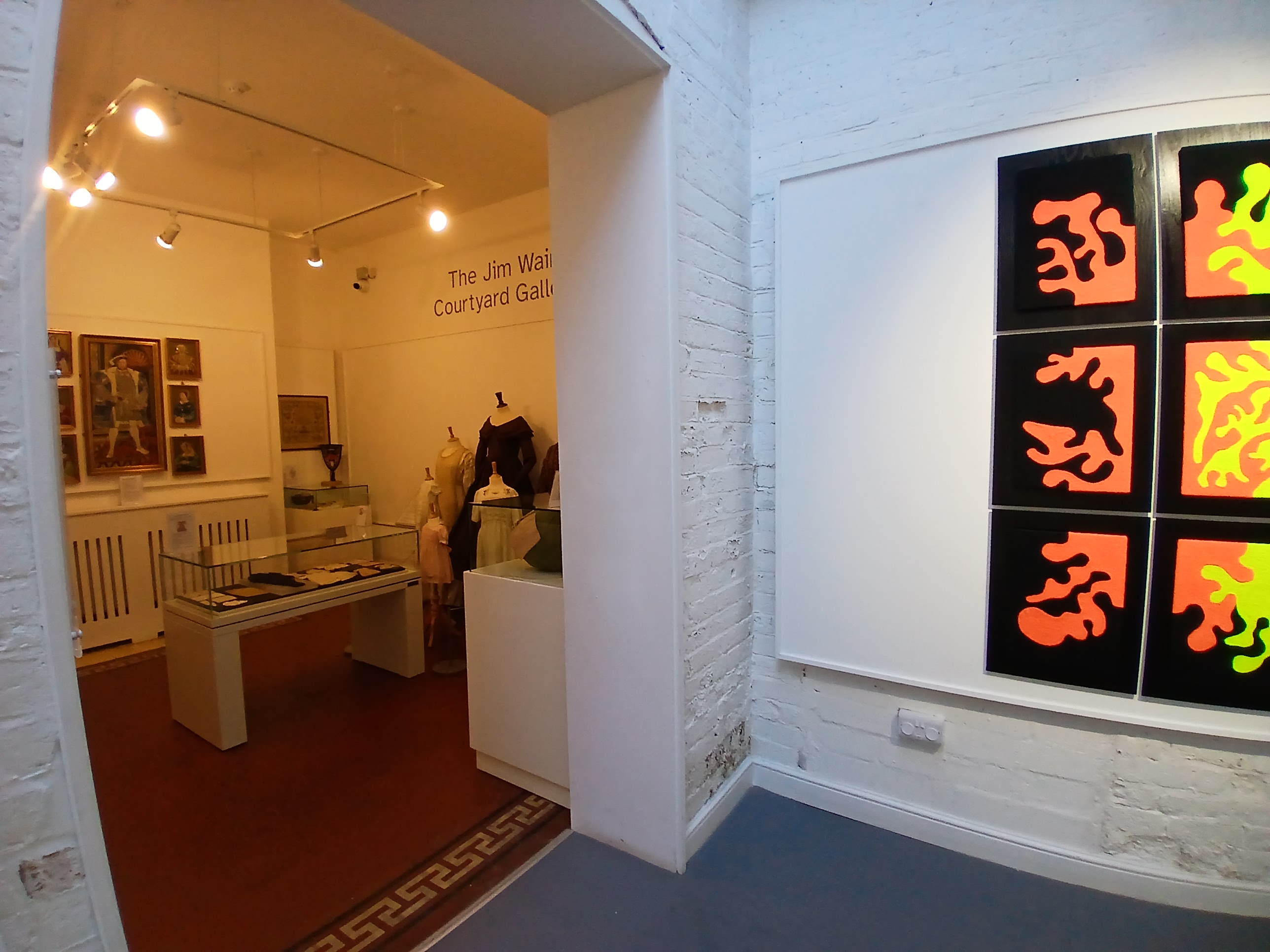 Looking through a large open doorway into a room with display cases, wall embroideries and mannequins. A brightly coloured artwork is on the right hand side.
