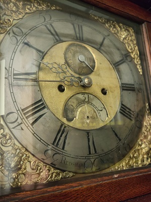 An ornamental clock face from a long case clock, mostly in yellow brass but with the numbers shining in silver