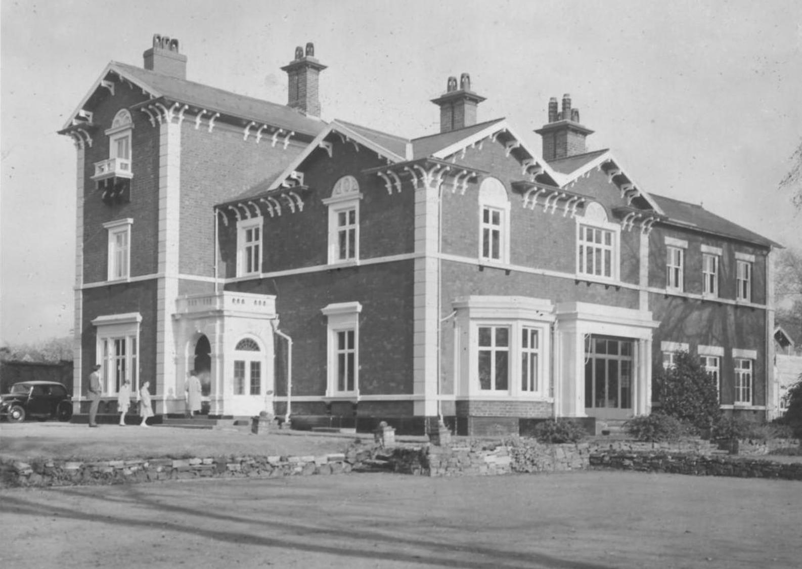 A black and white photograph of the Brampton Museum with a group of four people entering through an historic front door and just one car parked outside