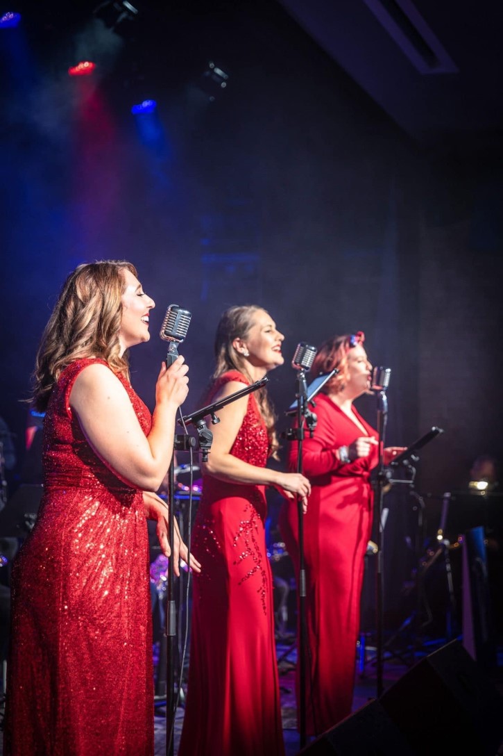 A trio of female singers in long, red sequined dresses on an indoor stage with vintage style microphones