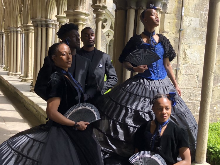 A group of black dancers pose in Victorian costume by an historic colonnade