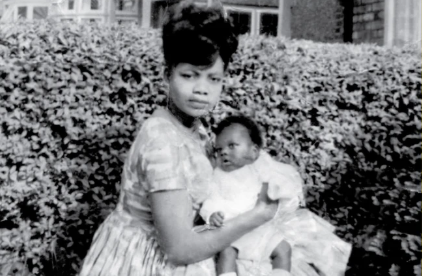 A black and white photograph of a black woman holding a baby in a garden