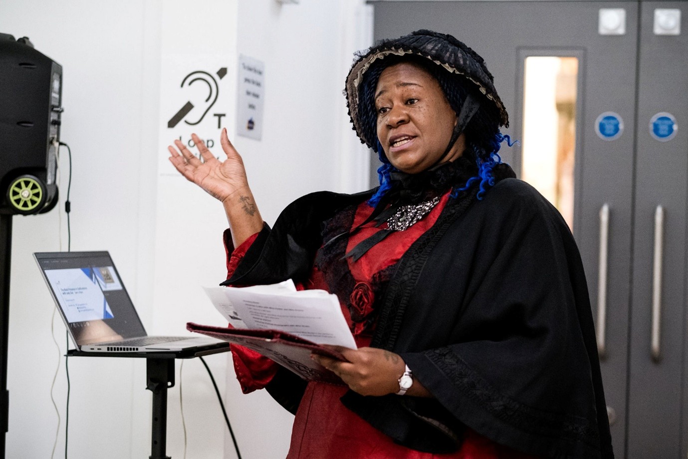 A black woman in a Victorian dress and bonnet gestures at a display screen to the right