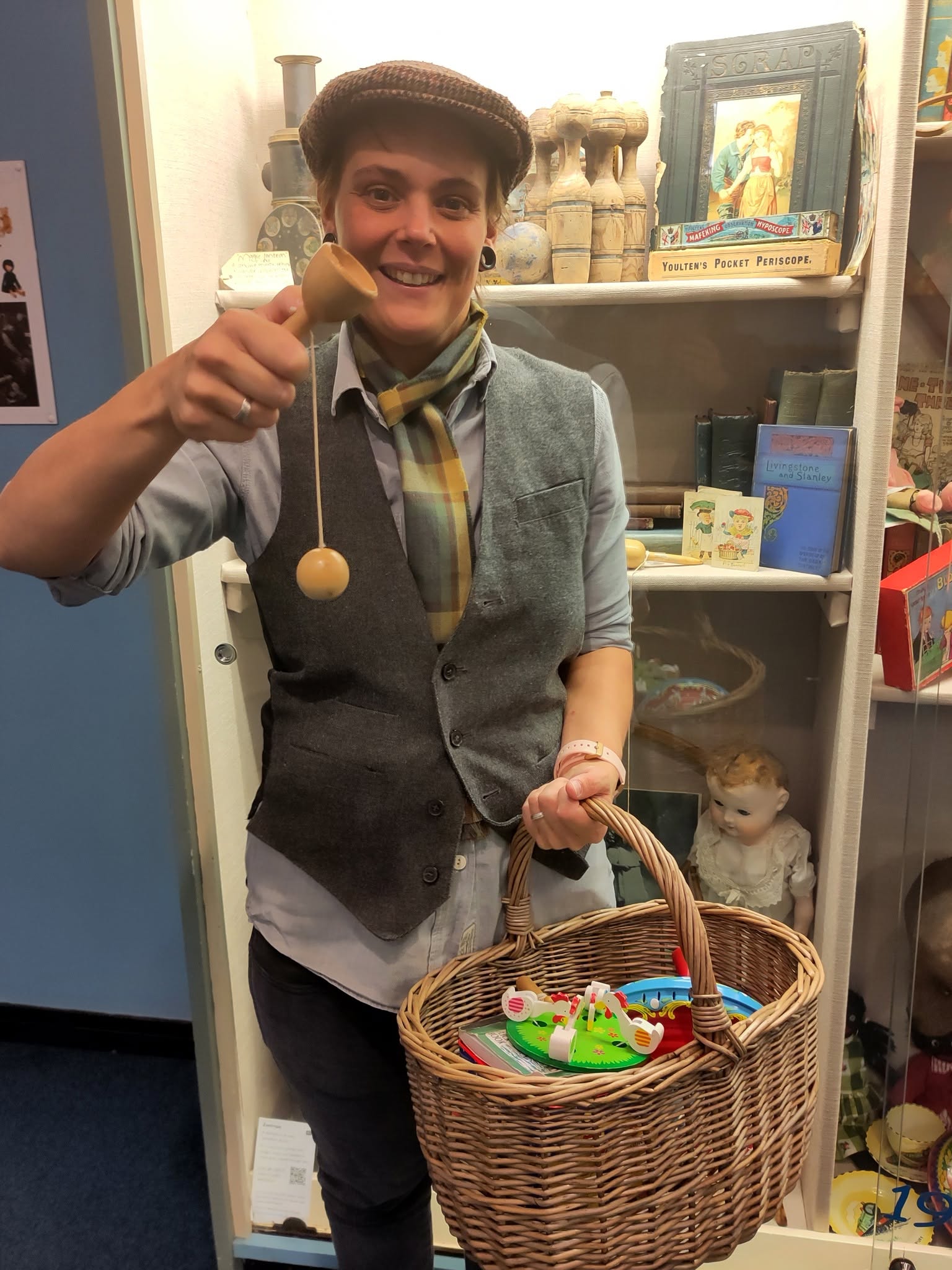 A person dressed as a Victoria schoolboy with flat cap and jacket holds a basked of toys in one hand and a cup and ball in the other
