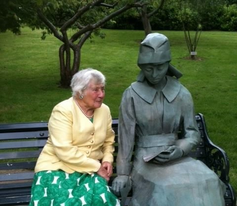 A metal statue of a woman on a park bench, wearing a WW1 uniform and sadly reading a letter. Next to here is an older woman, Baroness Sirley Williams, daughter of Vera Brittain.
