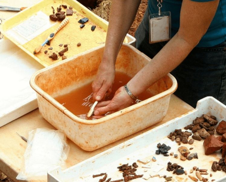 A person on an archaeological site washes small finds with a toothbrush in a plastic bowl. The bowl water is discoloured brown with soil. To the side is a tray of small, clean objects sorted into pottery shards, stone, bones etc.